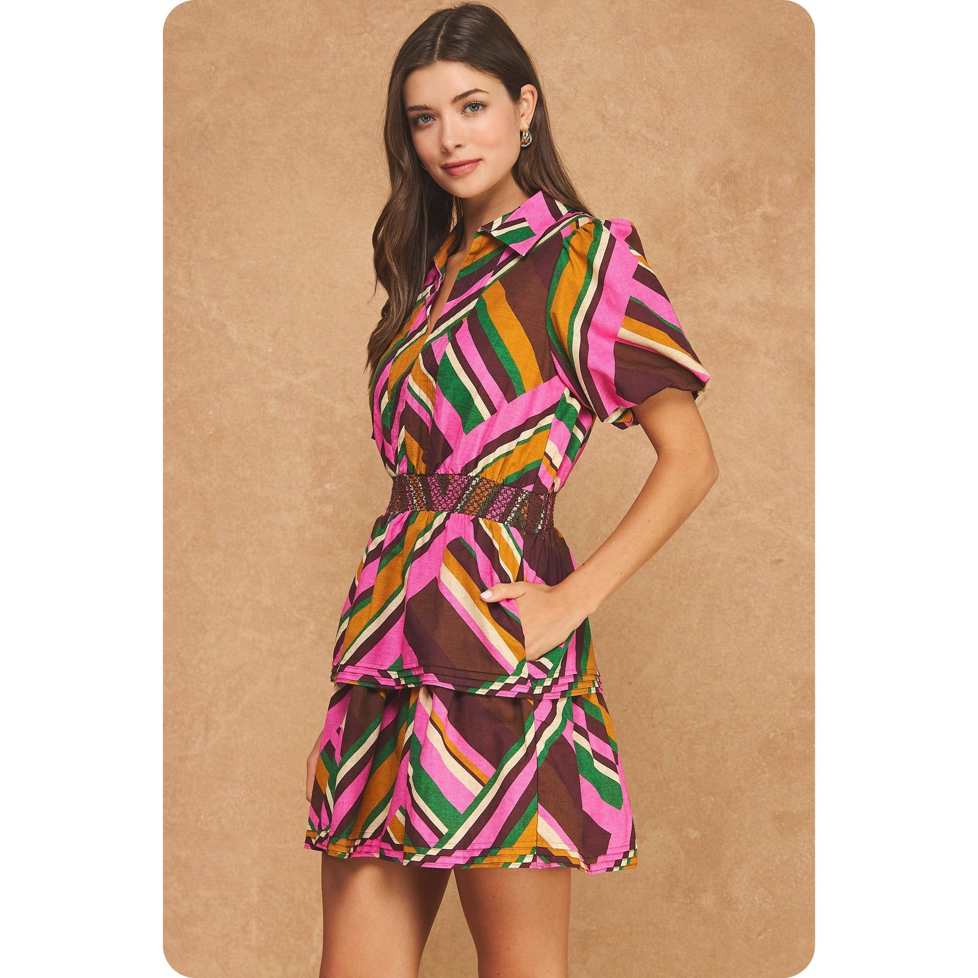 A woman modeling a short puff-sleeve mini dress with a tiered skirt and a geometric striped pattern in pink, brown, green, orange, and white, set against a textured beige background.