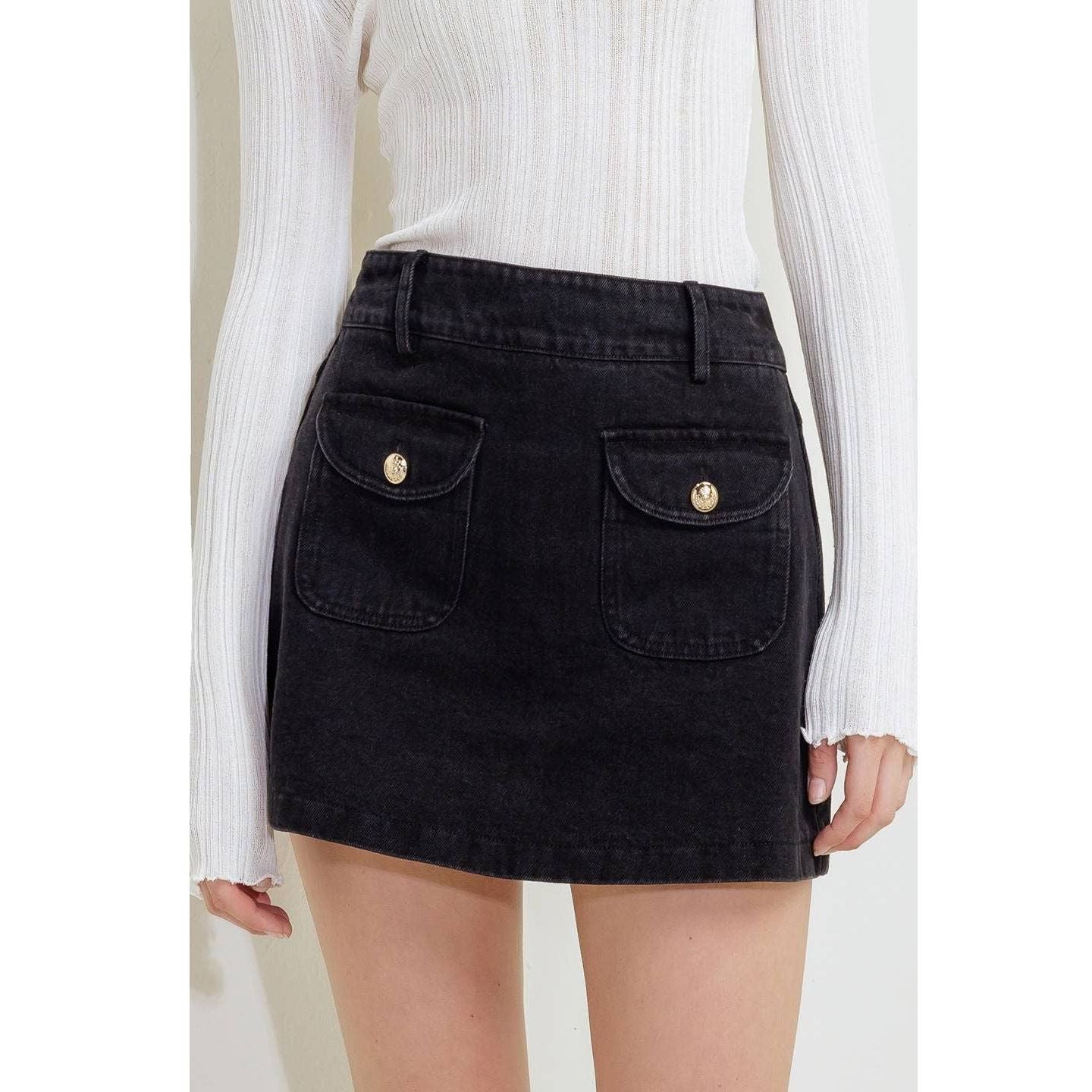 A model wearing a black denim mini skirt with two front flap pockets featuring gold-tone buttons, paired with a white ribbed long-sleeve top.