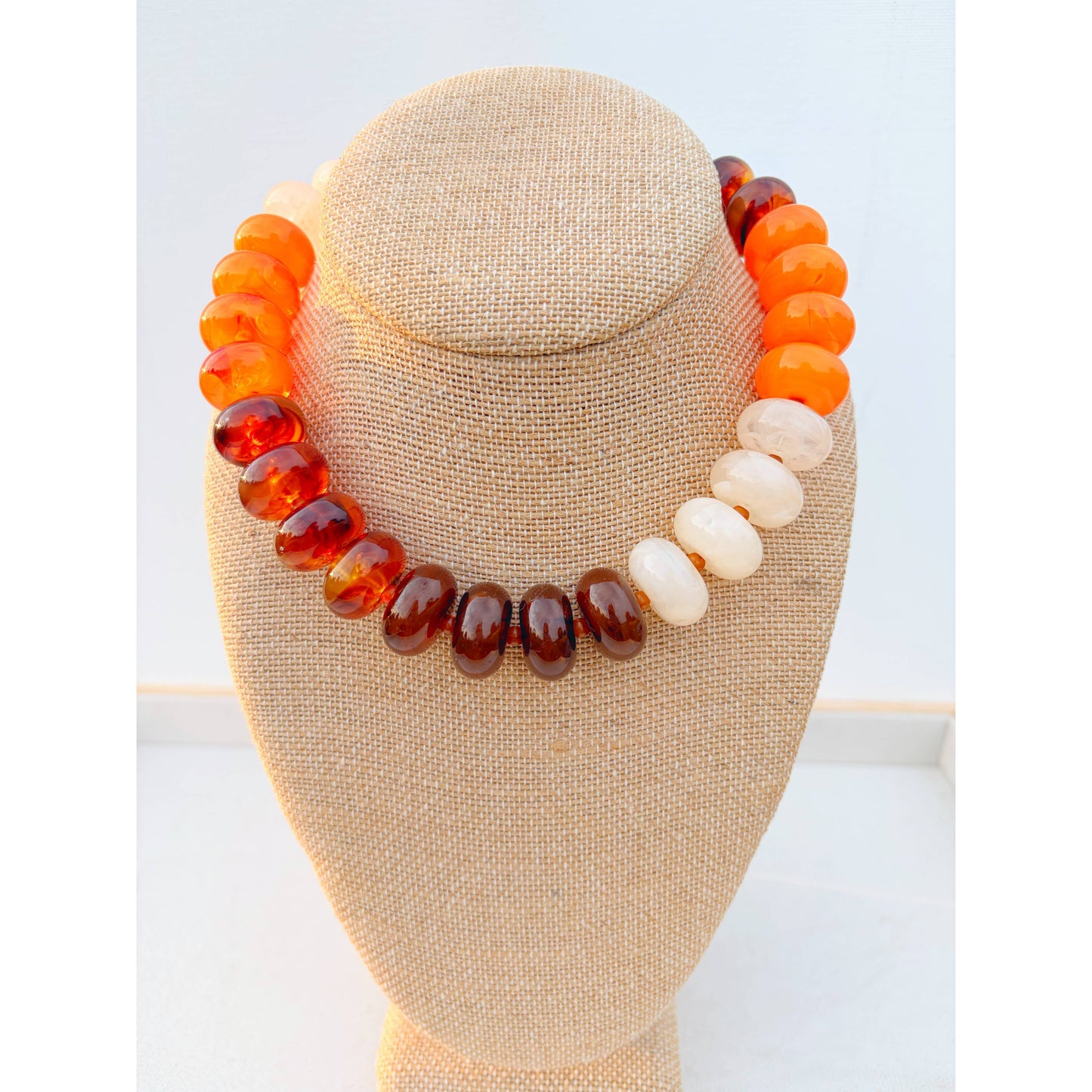 A chunky beaded necklace displayed on a beige fabric jewelry bust. The necklace features translucent orange and amber beads on one side, smooth white beads in the center, and polished dark brown beads on the other side, set against a clean white background.