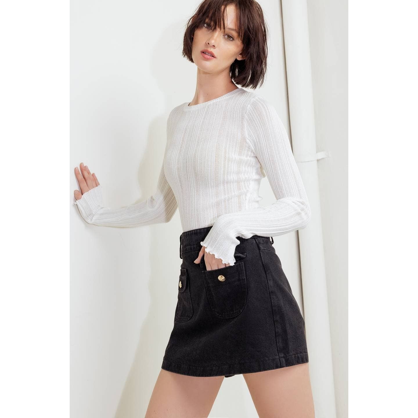 A woman wearing a white ribbed long-sleeve top with ruffled cuffs and a black denim mini skirt with front patch pockets, posing against a white wall.
