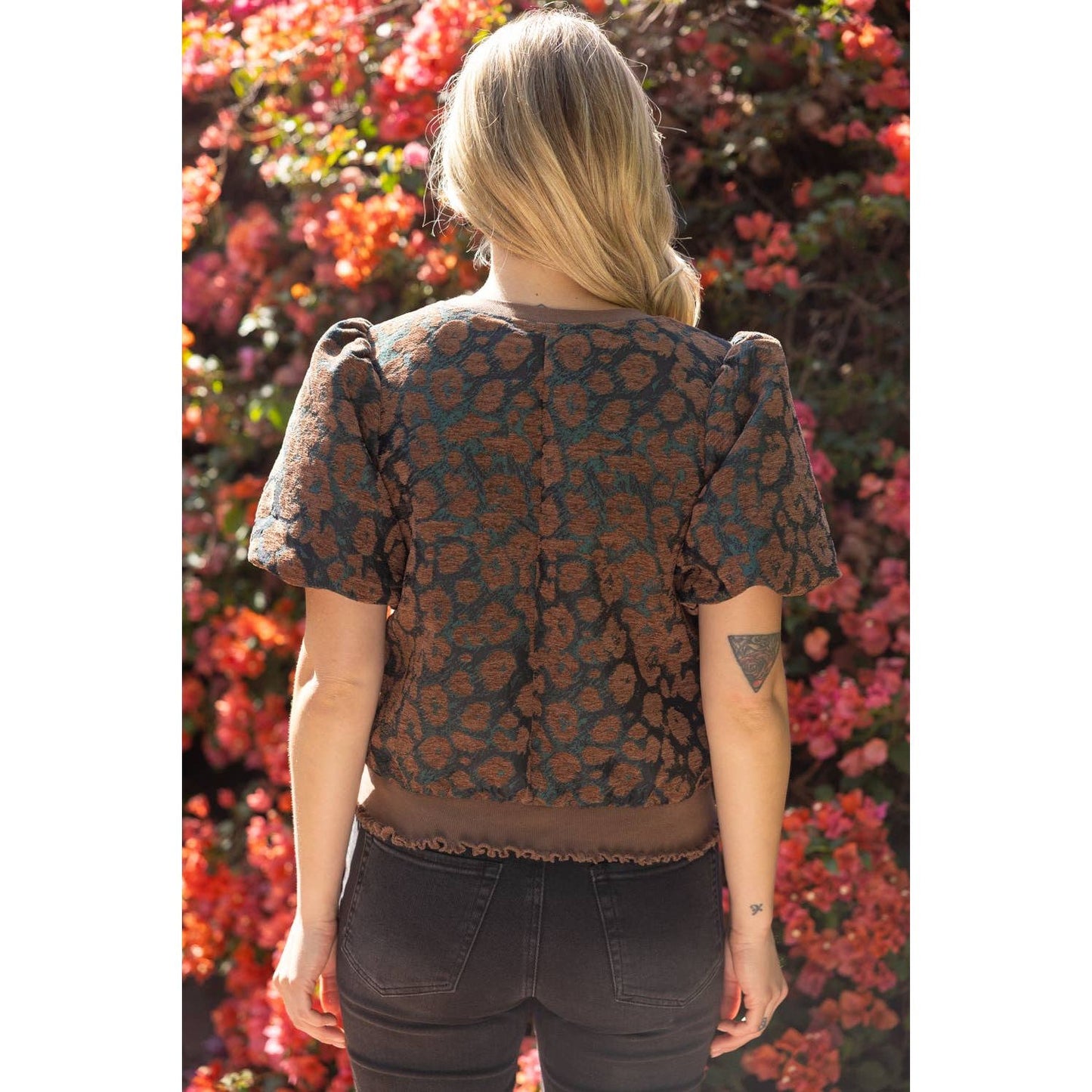 A woman viewed from behind wearing a short-sleeve cropped top with puff sleeves and a brown floral jacquard pattern on a dark background, set against a background of blooming pink flowers.