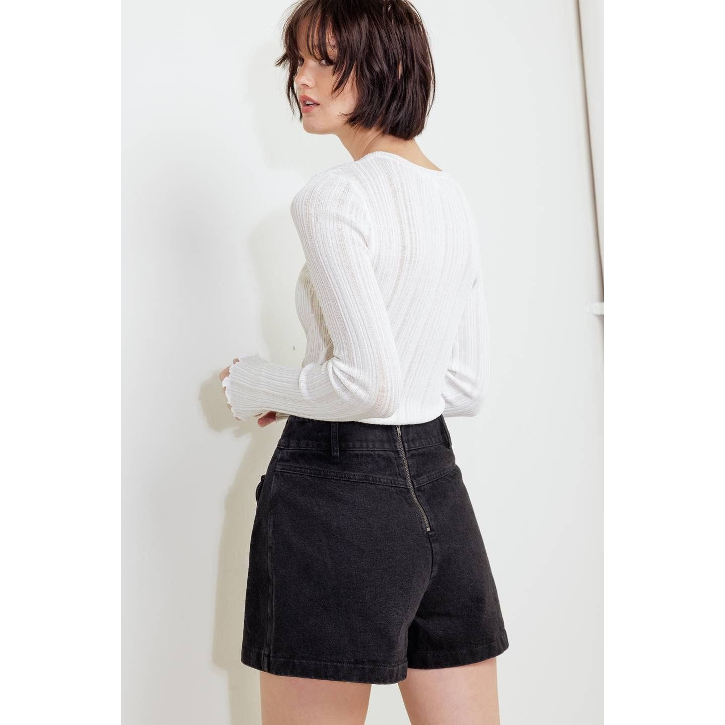 A woman wearing a white ribbed long-sleeve top and black denim shorts, photographed from the back against a plain light background.